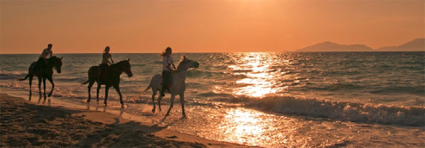 αναβάτες σε άλογα κάνουν βόλτα σε παραλία με τη δύση του ηλίου