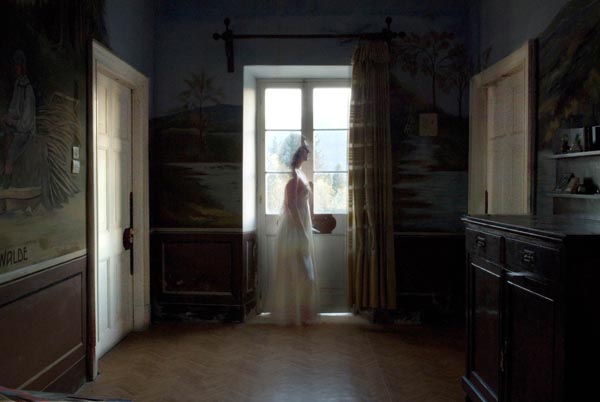 γυναικεία φιγούρα με άσπρο μακρύ φόρεμα προχωράει μπροστά απο ενα παράθυρο