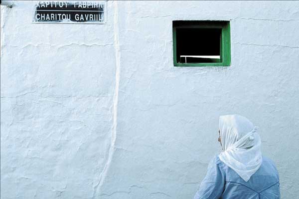 γυναίκα με μαντήλα μπροστά σε ένα κτήριο με ένα μικρό παράθυρο και το όνομα της οδού Χαρίτου Γαβριήλ