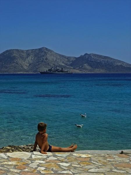 αγόρι κάθεται στο πλακόστροτο με μαγίο και κοιτάει δύο γλάρους στη θάλασσα / στο βάθος ένα καράβι και απο πίσω βουνό