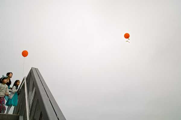 μια κοπέλα και δύο μικρά κορίτσια κρατούν ένα πορτοκαλί μπαλόνι και κοιτούν ένα άλλο πορτοκαλί μπαλόνι που έχει ξεφύγει