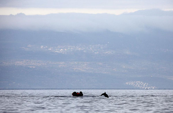 μία βάρκα με μετανάστες στη θάλασσα και δίπλα τους ένα δελφίνι που πηδά απο τη θάλασσα