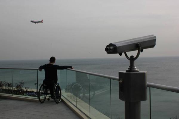 άντρας σε αναπηρικό αμαξίδιο κοιτάζει τη θάλασσα στο βάθος ένα αεροπλάνο και στα δεξιά της φωτογραφίας ένα τηλεσκόπιο