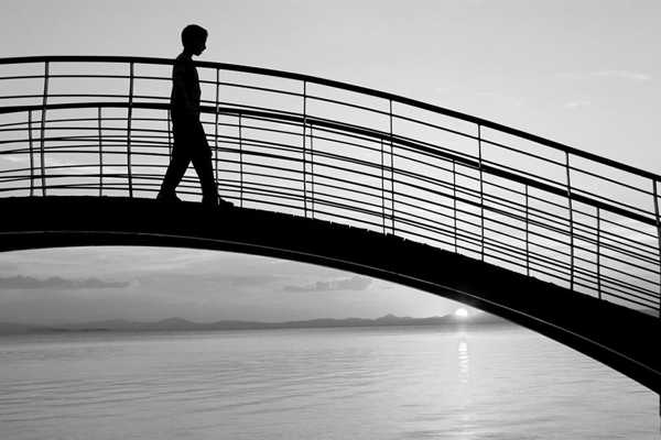 ασπρόμαυρη φωτογραφία με τη σιλουέτα ενός αγορίου πάνω σε μια γέφυρα και στο πισω μέρος θάλασσα