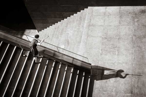 ασπρόμαυρη φωτογραφία γυναίκας που καθαρίζει σκάλες και η σκιά της φαίνεται σα να καρφώνει κάποιον με κοντάρι