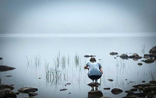 φωτογραφία μίας κοπέλας που στέκεται πάνω σε μία μικρή πέτρα και γονατιστή κοιτά μία λίμνη
