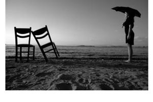 γυναίκα με μαύρη ομπρέλα δίπλα από 2 ξύλινες καρέκλες στη παραλία
