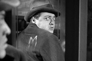 άνδρας απο την ταινία «Μ» κοιτά πίσω του φοβισμένος, φοράει καπέλο και κοστούμι και στην πλάτη του είναι σημειωμένο το γράμμα «Μ»