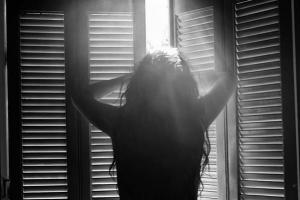 ασπρόμαυρη φωτογραφία / κοπέλα στέκεται μπροστά σε παράθυρο με μισάνοιχτα παντζούρια απο τα οποία περνάνε μέσα ηλιαχτίδες