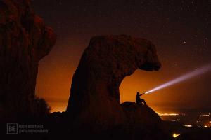 ένας άνδρας πάνω σε ένα βράχο φωτίζει με φακό τον κόκκινο ουρανό