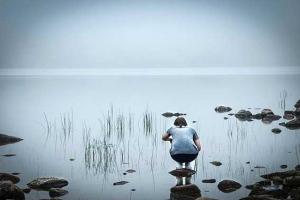 φωτογραφία μίας κοπέλας που στέκεται πάνω σε μία μικρή πέτρα και γονατιστή κοιτά μία λίμνη